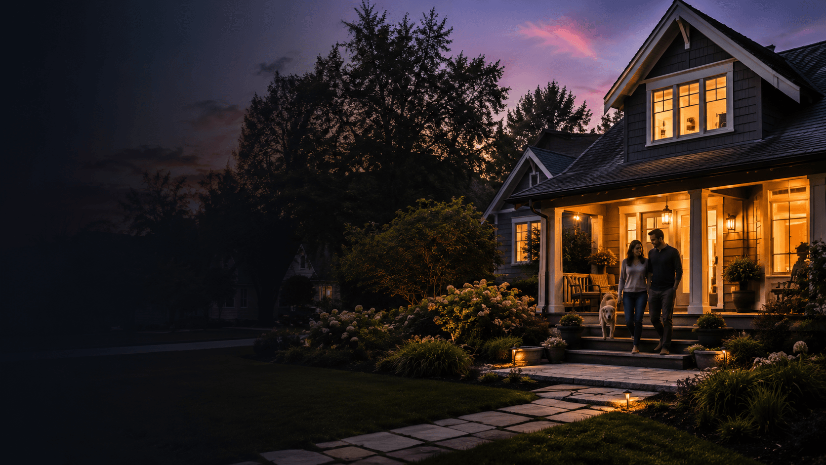 A couple and their dog on the porch of a warm craftsman home at twilight, its windows glowing